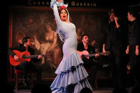 La mejor bailaora de flamenco, con un vestido azul voluminoso, cautiva en el escenario mientras dos guitarristas la acompañan.