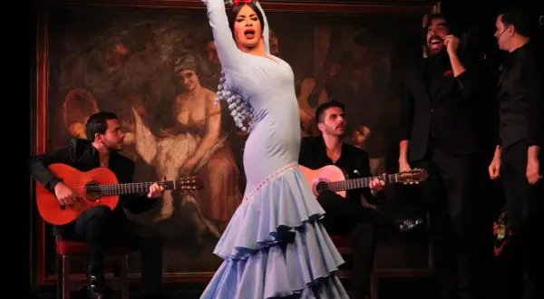 La mejor bailaora de flamenco, con un vestido azul voluminoso, cautiva en el escenario mientras dos guitarristas la acompañan.