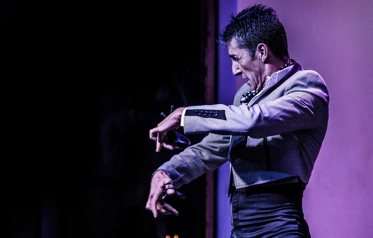 Un hombre con traje a cuadros realiza un apasionado flamenco baile, con las manos extendidas y castañuelas.