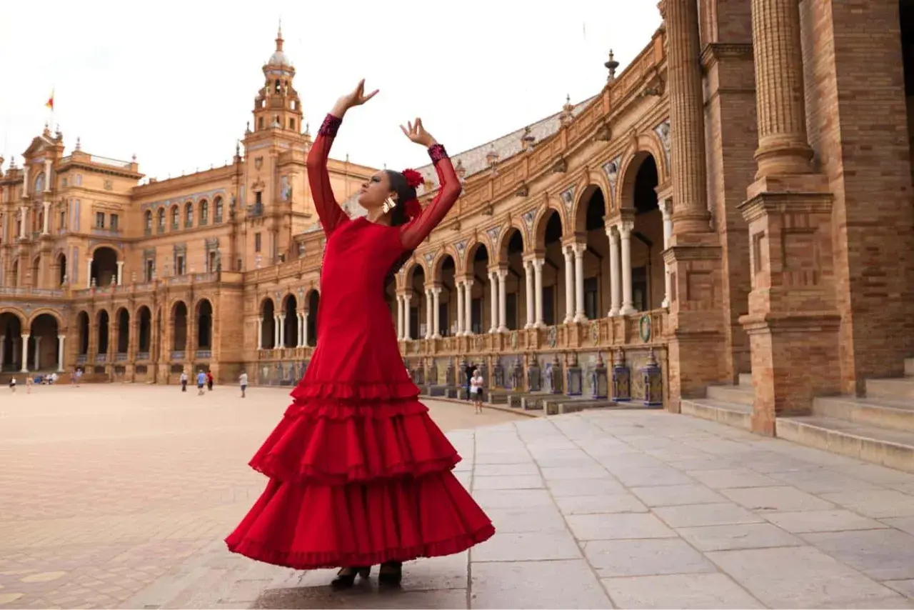 Bailarina con vestido rojo voluminoso, brazos en alto, ejecutando sevillanas lentas frente a un edificio histórico con arcos.