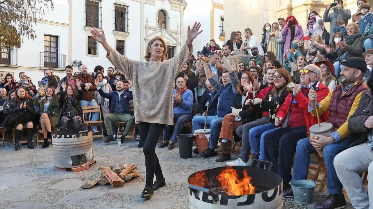 Una mujer baila animada mientras una multitud celebra las rumbas navide&ntilde;as. El fuego crepita en un barril, creando un ambiente festivo.