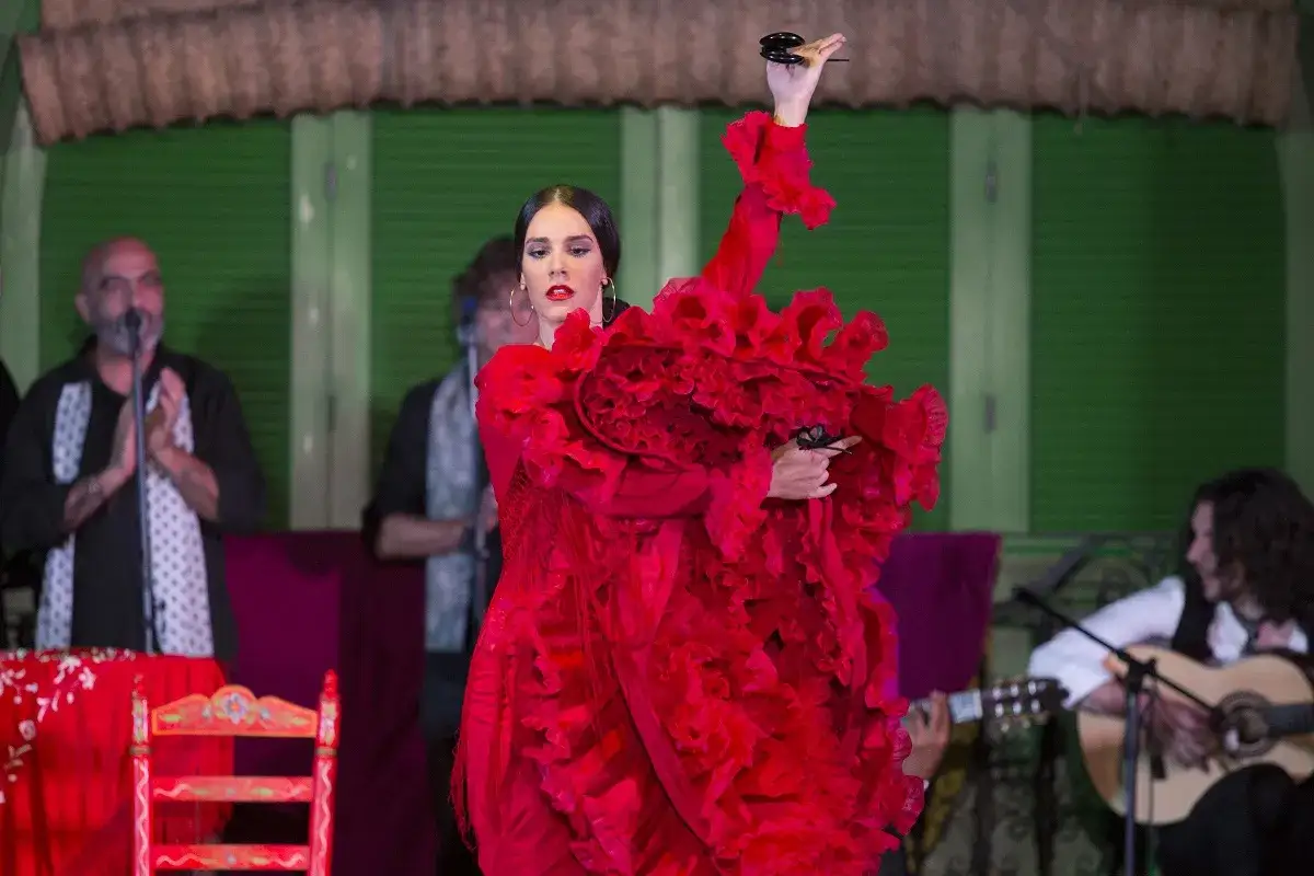Bailaora de flamenco cl&aacute;sico con vestido rojo voluminoso, casta&ntilde;uelas en mano, acompa&ntilde;ada por guitarrista y cantaor.