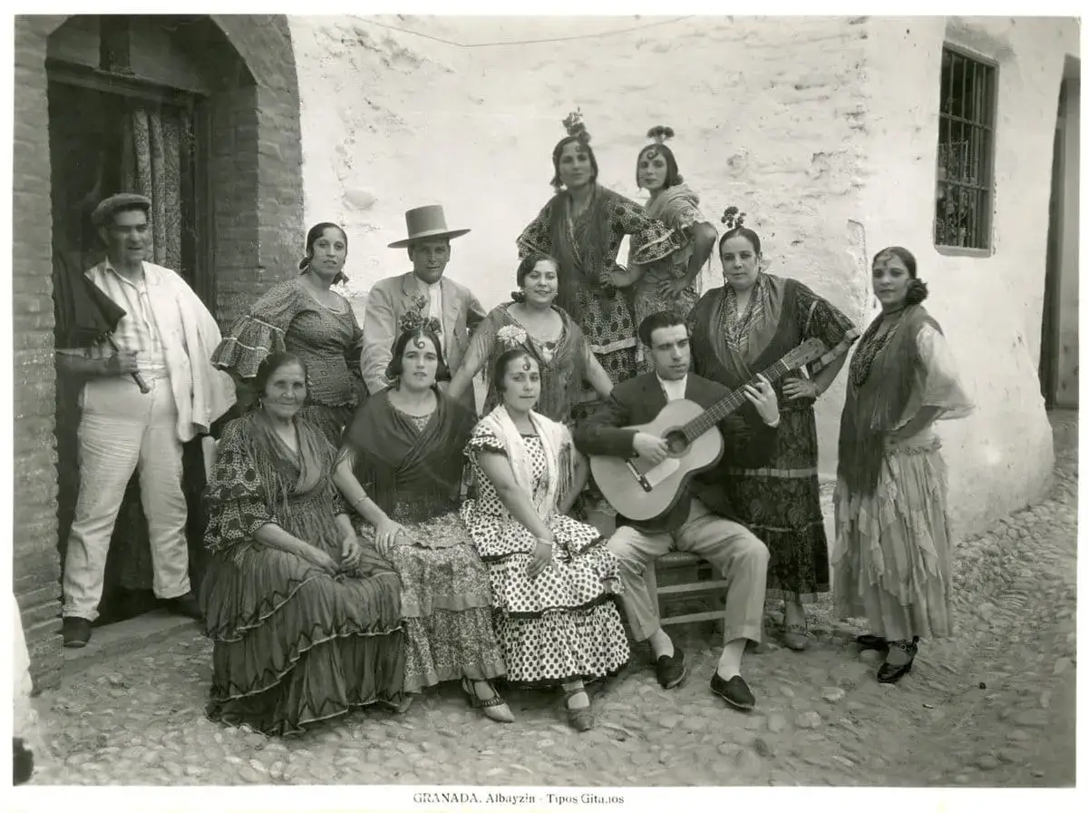 Grupo de personas vestidas con trajes flamencos, algunos con mantones y peinetas. Un hombre toca la guitarra. Los nombres flamencos de estas figuras resuenan en la tradici&oacute;n.