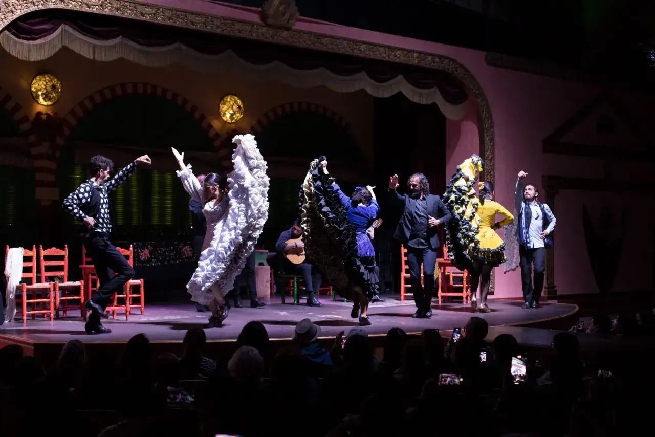 Artistas bailan al ritmo de canciones de sevillanas en un escenario con sillas rojas y luces c&aacute;lidas.