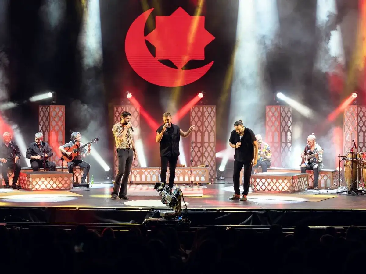 Artistas en un escenario con un gran logo rojo de luna y estrella. Un grupo interpreta música con guitarras y percusión, mientras tres cantantes lideran la actuación. El fondo flamenco baile crea una atmósfera vibrante.