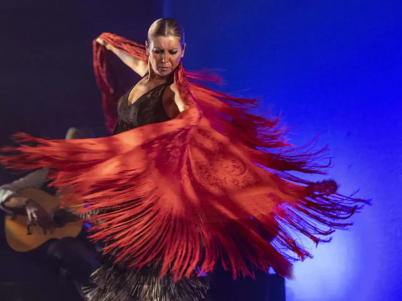 Sara Baras joven, con un mant&oacute;n rojo vibrante, baila flamenco con pasi&oacute;n. Un guitarrista la acompa&ntilde;a en el escenario.
