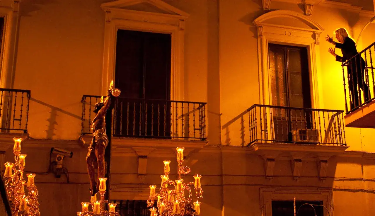 Procesi&oacute;n nocturna con figura de Cristo y candelabros iluminados. Una mujer observa desde un balc&oacute;n, evocando las saetas populares.