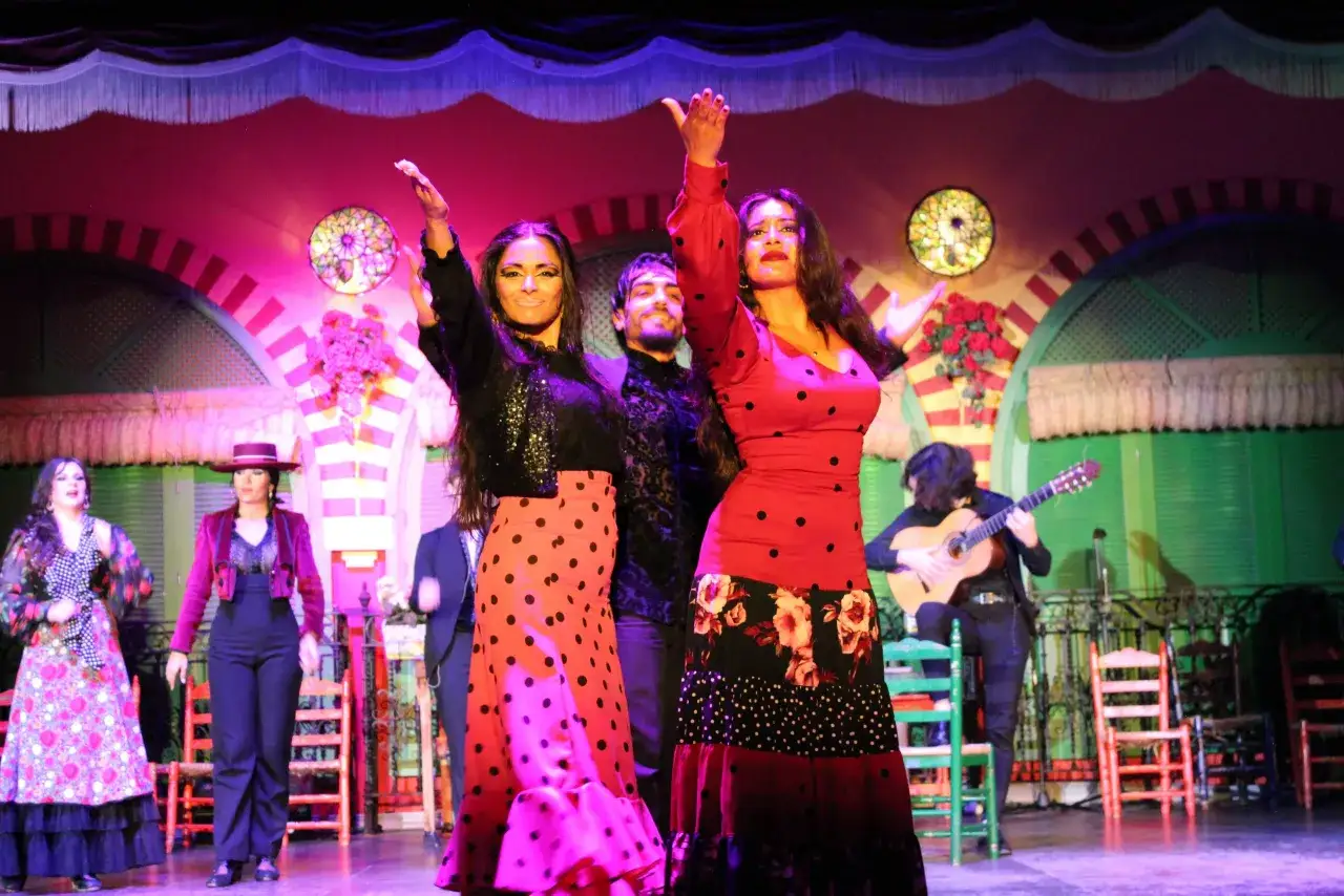 Artistas de flamenco en un escenario vibrante, con dos mujeres bailando al frente y un guitarrista al fondo. Un cante de ida y vuelta lleno de pasi&oacute;n.