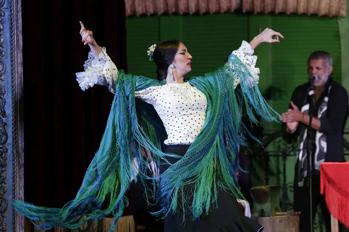 Bailarina flamenca con mant&oacute;n verde, interpretando apasionadas canciones flamencas de amor, mientras un hombre aplaude al fondo.
